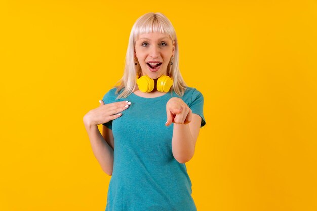 Pointing at camera with headphones blonde caucasian girl in studio on yellow background