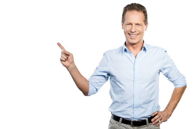 Pointing copy space. Happy young man in shirt looking at camera and pointing away while standing against white background