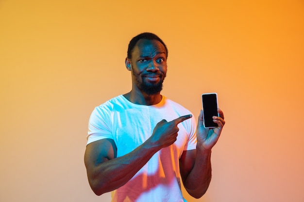 Pointing on phone with blank screen. African-american man's modern portrait on gradient orange studio wall