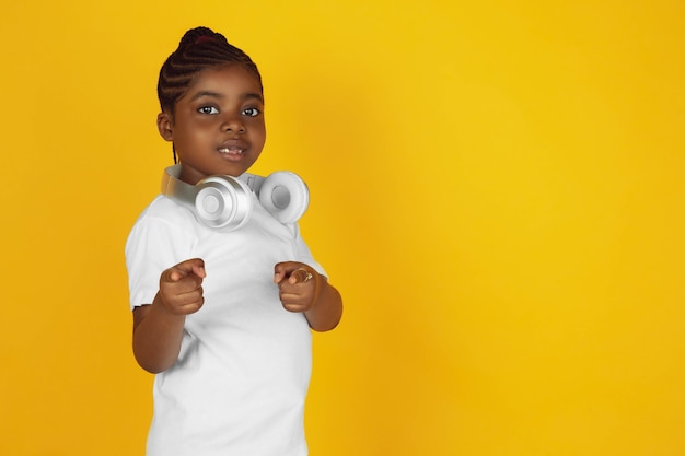 Pointing, putting on headphones. little african-american girl's portrait on yellow studio background. cheerful kid. concept of human emotions, facial expression, sales, ad. copyspace. looks cute