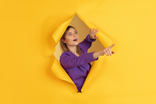 Pointing, showing. cheerful caucasian young woman poses in torn yellow paper background, emotional and expressive. breaking on, breakthrought. concept of human emotions, facial expression, sales, ad