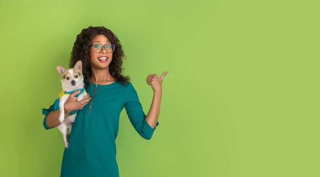 Pointing at side. African-american young woman's portrait isolated on green studio background. Beautiful female model with little doggy. Concept of human emotions, facial expression, sales, ad.