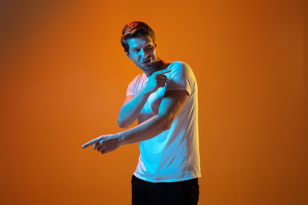 Pointing at side. Caucasian young man's portrait on gradient orange studio background in neon light. Beautiful male model. Concept of human emotions, facial expression, sales, ad. Copyspace.