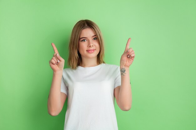 Pointing up. Caucasian young woman's portrait isolated on green studio wall.