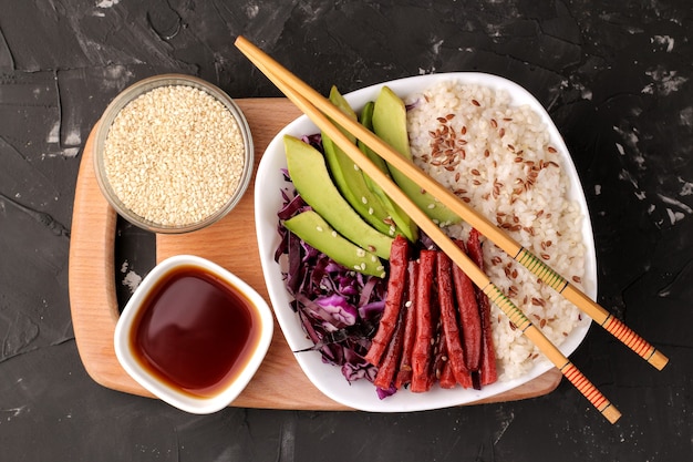 Poke bowl Hawaiiaans eten. een bord rijst, zalm, avocado, kool en kaas. in de buurt van skunk en sojasaus op een zwarte achtergrond. bovenaanzicht