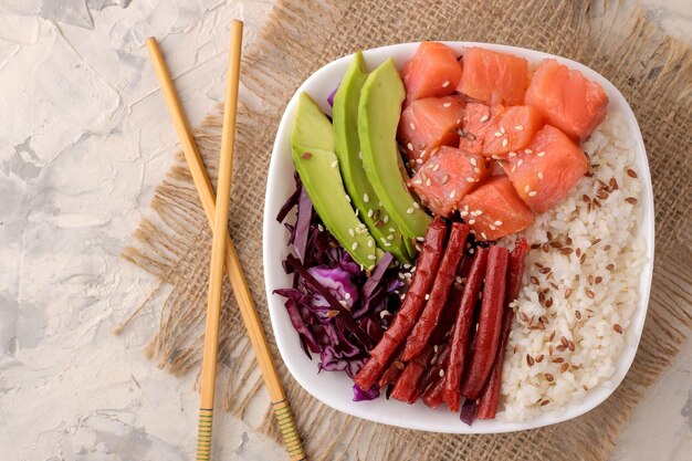 Poke bowl Hawaiian food. a plate with rice, salmon, avocado, cabbage and cheese on a light background with chopsticks. view from above