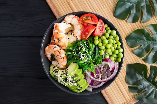 Poke bowl with red shrimps and vegetables in the dark bowl on the tropical background.Top view.Closeup.Copy space.