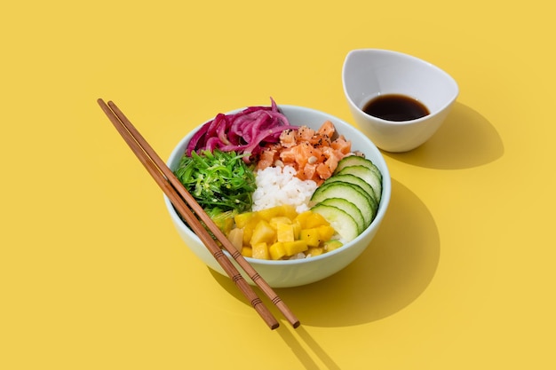 ご飯サーモンキュウリマンゴーオニオンワカメ サラダ ケシの実と黄色の背景に分離されたヒマワリの種とボウルを突く xA