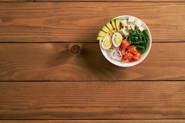 Photo poke bowl with salmon, avocado, cheese, eggs sprinkled sesame isolated on wooden background flat lay