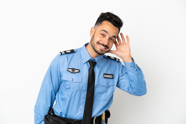 Police caucasian man isolated on white background listening to something by putting hand on the ear