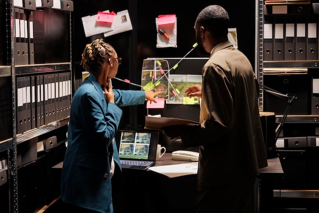 Police detective partners analyzing clues scheme on detective board. African american woman cop pointing at crime scene and suspect photos connection map, discussing insight with investigator