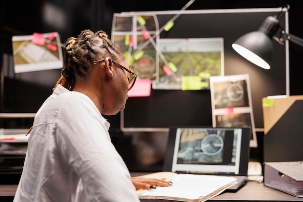 Police detective reading records in crime case file while working overtime in office. African american woman investigator checking infromation in law enforcement documentation