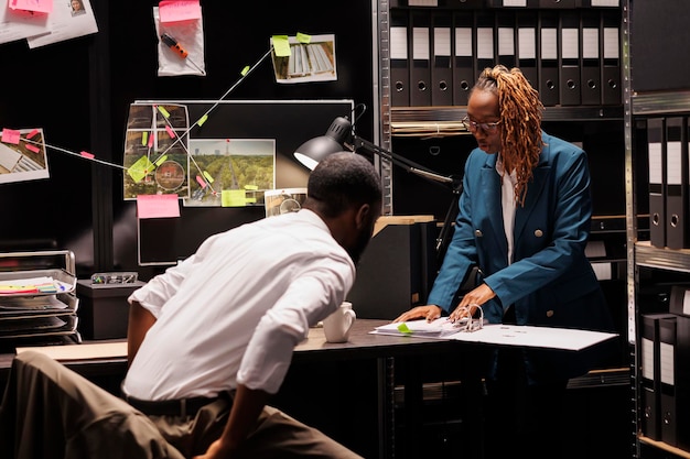 Police office secretary reading crime case file for detective. African american man investigator and woman assistant studying forensic expertise report and working at night time