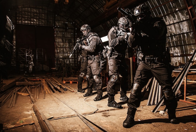 Police special operations anti-narcotics or counter-terrorism team, quick reaction tactical group in black uniforms and masks, armed with semi-automatic rifles moving with caution in abandoned hangar