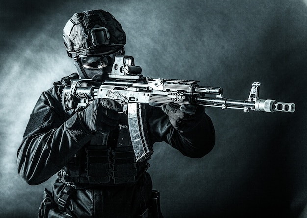 Police tactical team member, army special forces, private security company fighter in black uniform, helmet and mask aiming with collimator sight on assault rifle, toned in blue, low key studio shoot