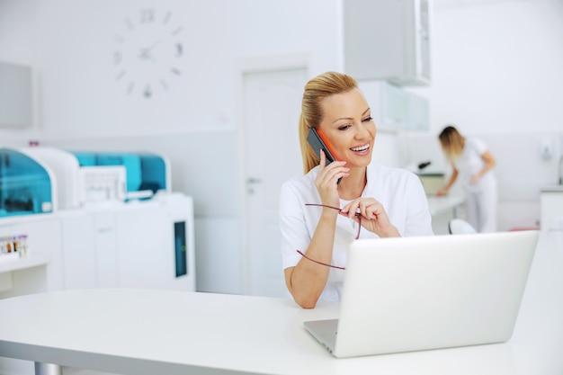 Polite attractive blond smiling lab assistant sitting in laboratory, having phone call with patient and looking at laptop with test results