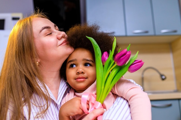 Poly-etnische moeder en haar dochter met afro-kapsel zitten in de keuken met een boeket tulpen en posrcard