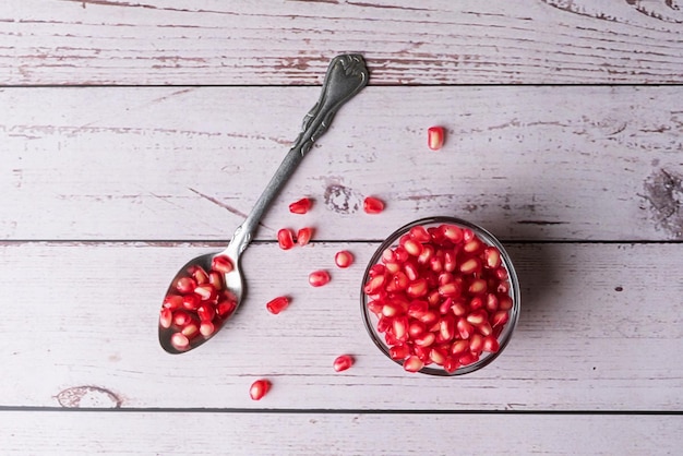 Pomegranate seeds in a glass bowl and a spoonful of red pomegranate seeds on a light wood background