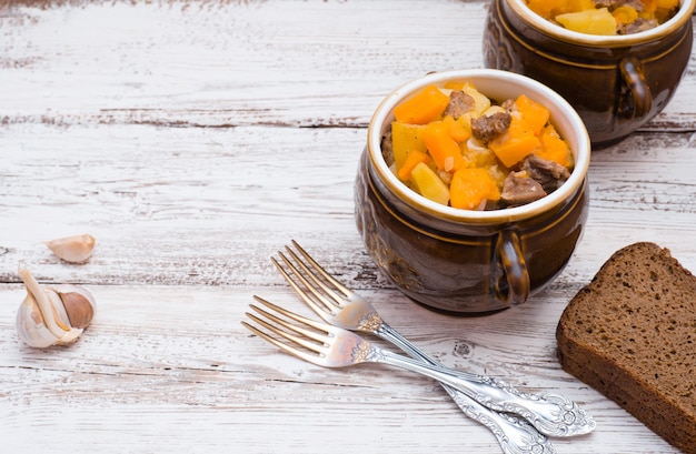 Pompoen, vlees, aardappelen en groenten gebakken in een keramische pot op een witte houten tafel.