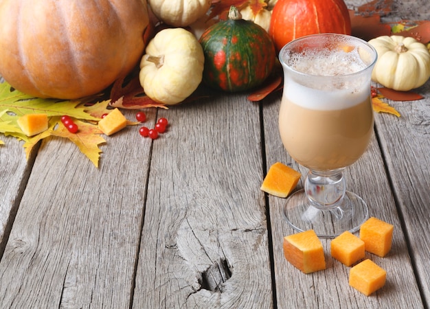 Pompoenkruid latte. glazen koffiekopje met romig schuim, gedroogde herfstbladeren, appels en kleine gele pompoenblokjes op rustiek hout. herfst warme dranken, seizoensgebonden aanbod concept