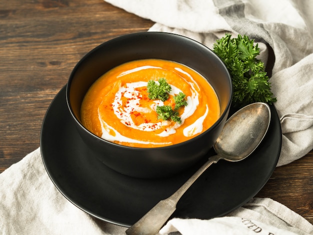 Pompoensoep met room, droge paprika en verse peterselie in een zwarte kom op een keukenhanddoek op een houten tafel