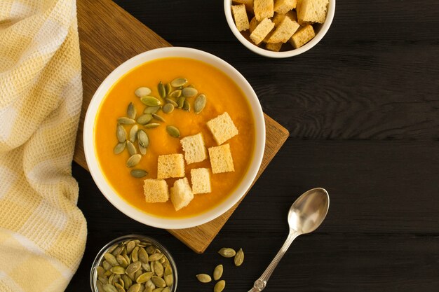 Pompoensoep met zaden en crackers in de witte kom op de bruine keuken boord op de zwarte houten achtergrond