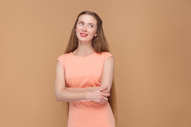 Photo pondering, dreaming beautiful woman looking away. portrait of emotional cute, beautiful woman with makeup and long hair in pink dress. indoor studio shot, isolated on light brown or beige background.