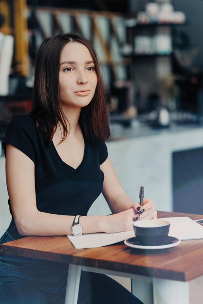 Pondering female student writes list to do in notepad thinks on future plans enjoys coffee break in cosy cafeteria notes information with pen concentrated into distance People leisure concept