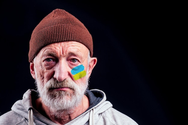 Poor caucasian old men with Ukraine flag painted on cheek in studio black background Russian versus Ukraine trade third world war disputes and anctions policy concept