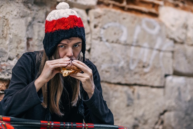 The poor dirty woman is eating a sandwich A hungry girl sits in dirty clothes wrapped in a blanket