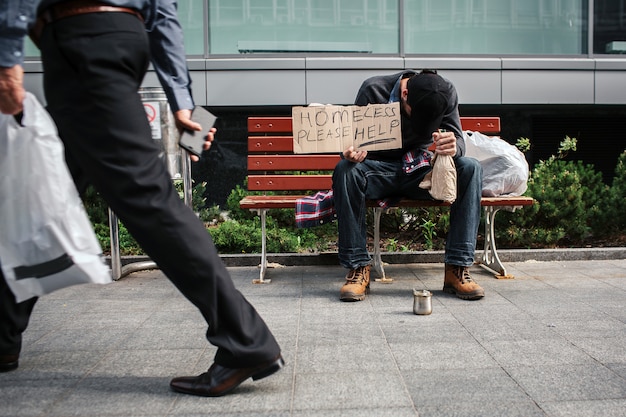 Poor and drunk man is sitting on bench and holding a cardboard which says homeless please help. He has put head on hand in which he has a bottle of drink. Guy is sleeping.