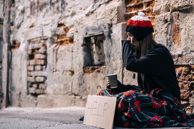 The poor girl is sitting on the street wrapped in a checkered blanket holding a glass in her hands asking for help crying wiping her eyes