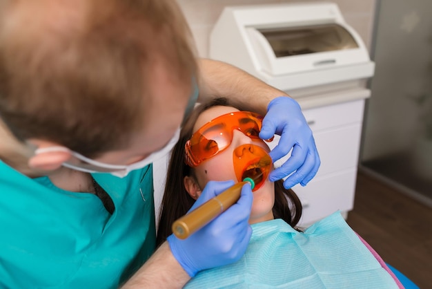 Poor girl in a patients bib and protective glasses in a dental clinic Dentist in blue latex gloves is removing her tooth with a help of a cheek retractor and forceps Closeup horizontal photo