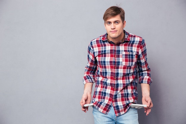 Poor handsome young man in checkered shirt and jeans showing empty pockets over grey wall