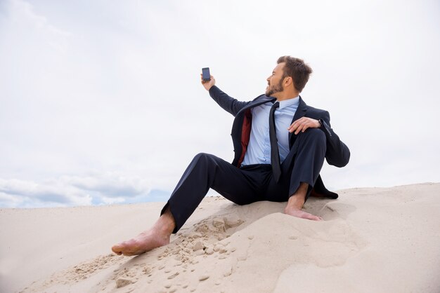 Poor signal. Frustrated young businessman searching for mobile phone signal while sitting on sand in desert