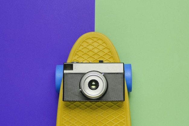 Pop art retro style still life. 80s. Cruiser board with retro camera on two-color paper background. Summer fun. Top view.