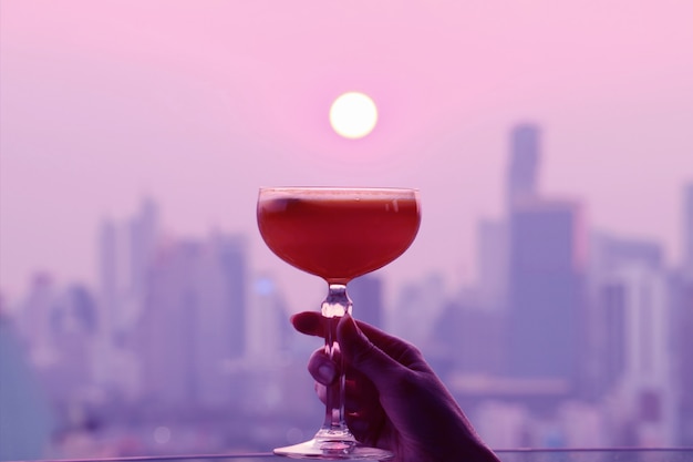 Pop art style pink and purple colored cocktail glass in hand with bright sun setting over the city