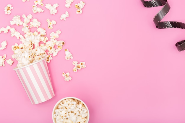 ピンクの背景のポップコーンバケツとフィルムストリップ。映画やテレビの背景。トップビューコピースペース