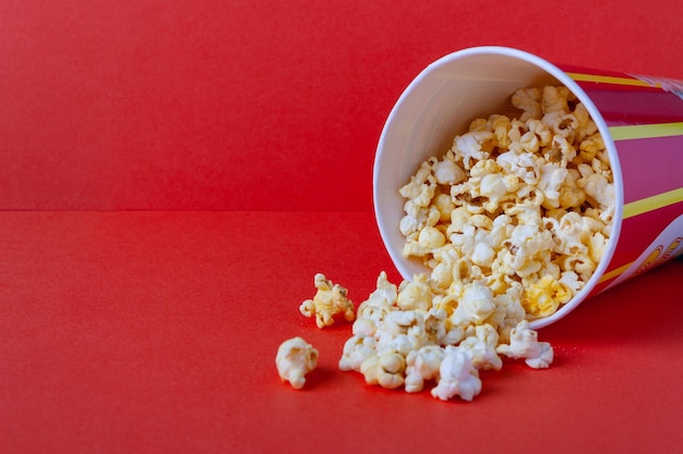 Popcorn spills out of a striped paper bucket on a red background copy space