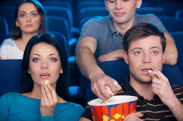 Popcorn stelen. opgewonden jong stel dat popcorn eet en film kijkt in de bioscoop terwijl de man van de achterbank hun popcorn steelt