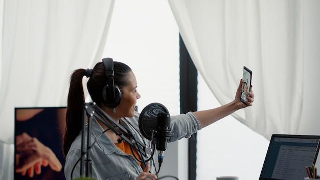 Populaire social media-beïnvloeder die selfie-foto maakt met smartphone tijdens het streamen van internetshow. Creatieve contentmaker zit in de thuisstudio terwijl hij foto's maakt met een mobiele telefoon met touchscreen.