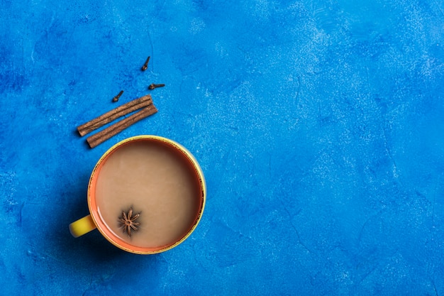 Popular Asian warming drink masala tea in a yellow cup with coriander, cinnamon and rose stem on a classic blue background.