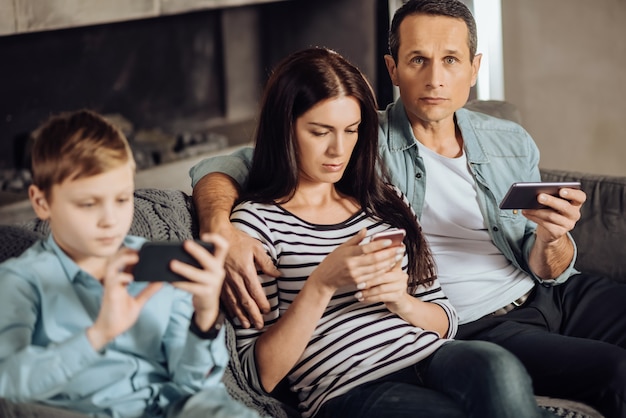 Popular pastime. Pleasant young man sitting on the couch next to his wife and son, hugging his wife and looking at camera while everyone being glued to their phones