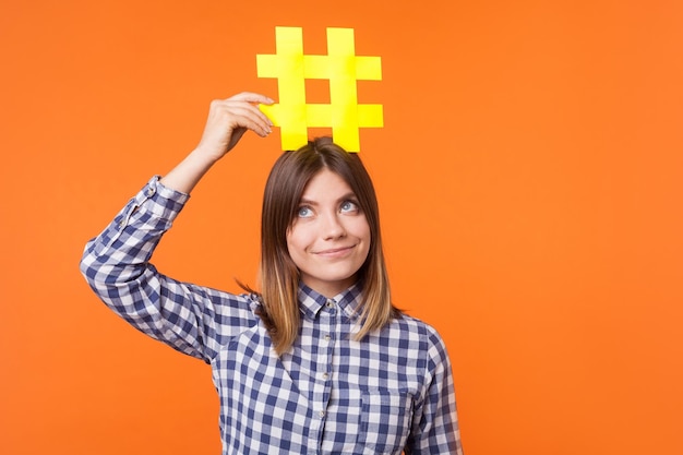 popularity in social media portrait of funny brunette woman wearing checkered casual shirt holding large big yellow hashtag sign on head as crown indoor studio shot isolated on orange background