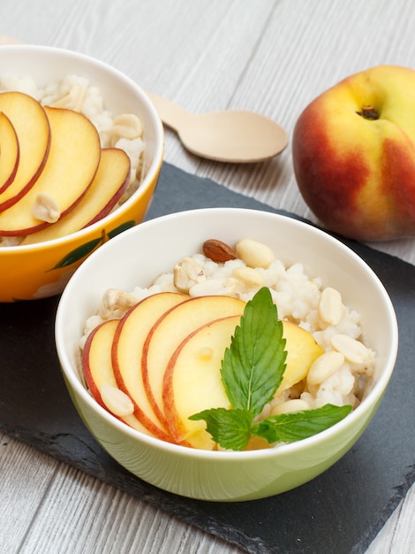 Porcelain bowls with sorghum porridge, peach, cashew nuts, almond and mint leaves, wooden spoon on a black stone board. Vegan gluten-free sorghum salad with fresh fruits. Top view.