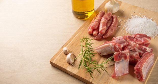 Pork ribs, sausage segments, rock salt, garlic, onion, rosemary and olive oil on a light rustic wood on a table, selective focus.