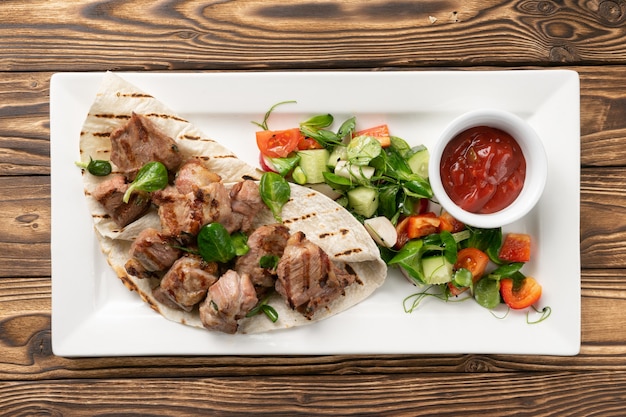 Pork shish kebab in lavash with ketchup and vegetable salad of cucumbers, tomatoes and bell peppers in a white ceramic plate