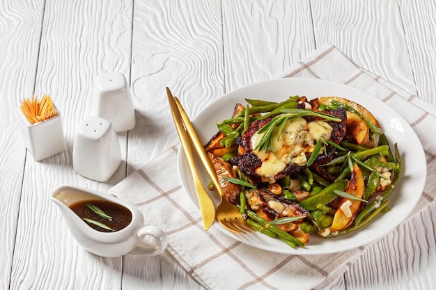 Pork steak topped with melted blue cheese on a pad of roasted\
sugar snap peas potato wedges green beans and garlic with dipping\
sauce in a gravy boat horizontal view