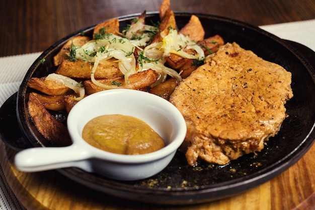 Pork steak with fried potatoes in a frying pan served with mustard sauce in a restaurant