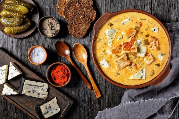 Pork stew with cream, pickles, and blue mold cheese cooked of lean pork fillet with smoked paprika and black pepper served on a clay pot with a wooden spoon on a dark wooden table, top view, close-up